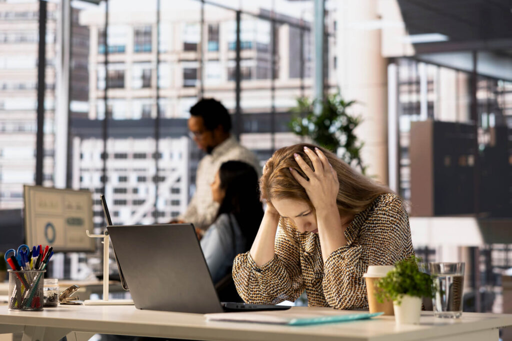 Stressed office worker sitting at desk with head in hands, laptop open, and colleagues working in the background